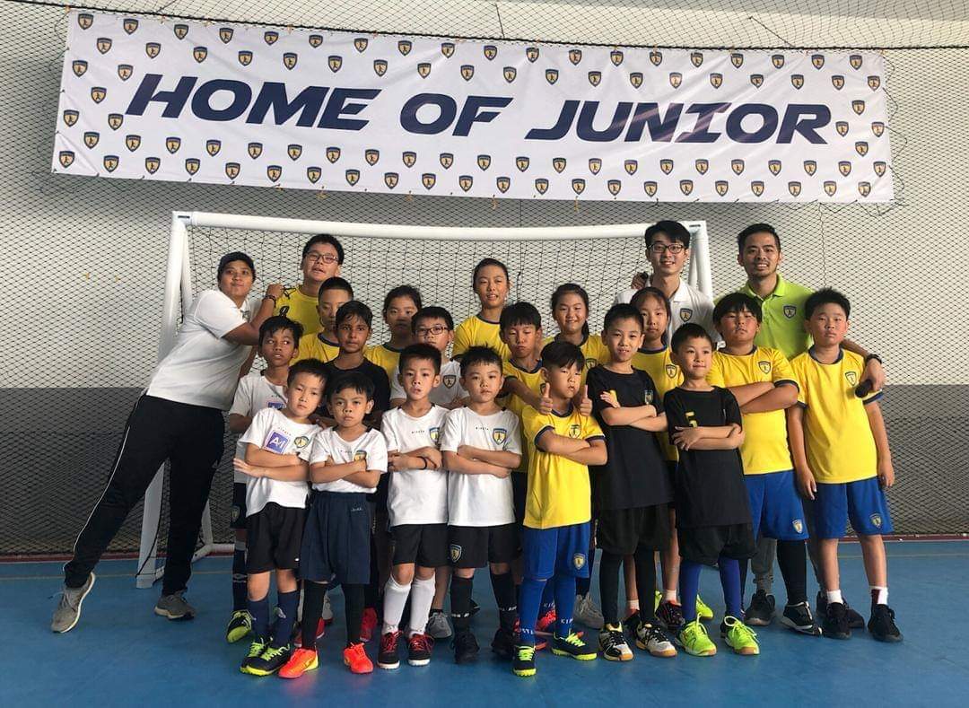 Players and coaches posing together in front of the Home of Junior banner at an indoor football court.