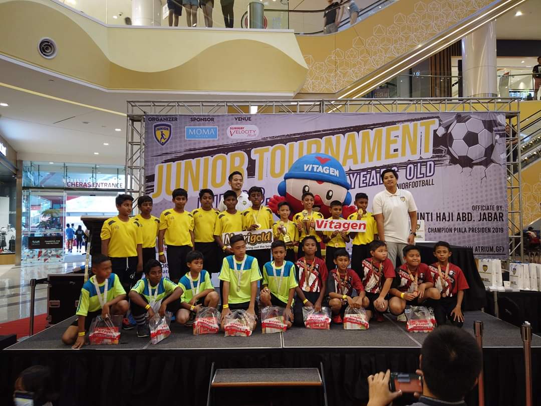 Young players and coaches gathered on stage with medals and prizes at a junior football tournament presentation.