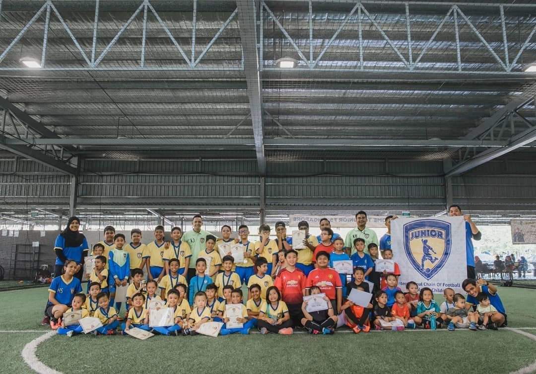 Players and coaches displaying certificates and medals beside an academy banner on an indoor pitch.